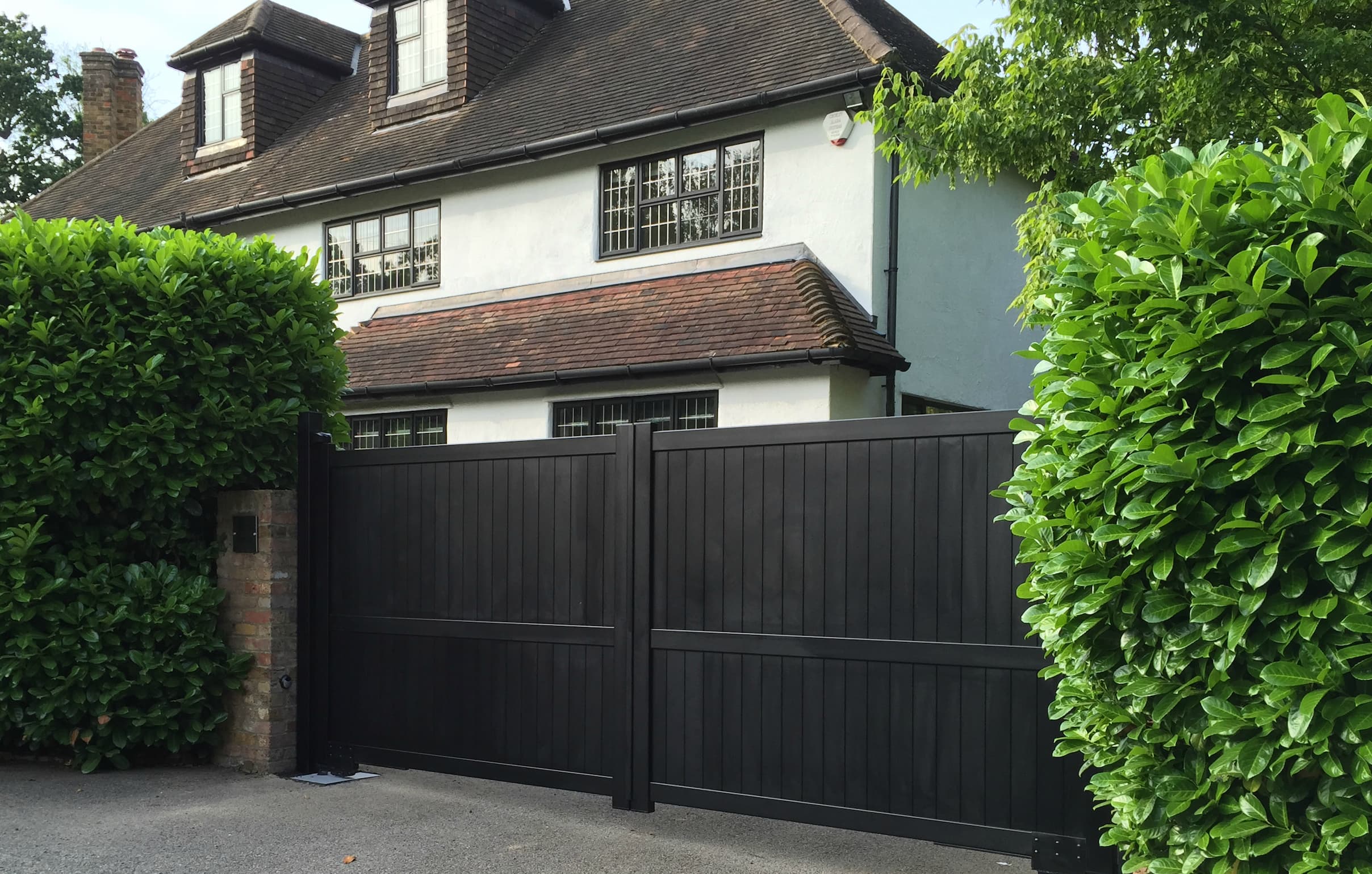 Traditional house with a dark brown aluminium gate, flanked by vibrant green shrubbery.